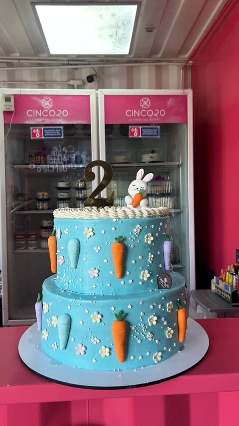 Two-tier turquoise birthday cake decorated with white flowers, carrots, and a bunny topper with number 2, displayed in a bakery window with pink branding