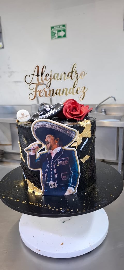 Decorative black cake with gold accents, a printed photo of a man in a sombrero, a red rose, and a golden Alejandro Fernandez name topper on a white cake stand