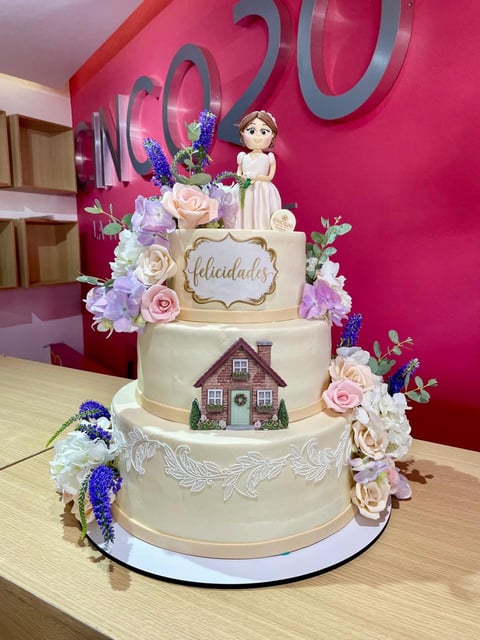 Three-tiered decorated cake with bride figurine, edible house design, purple and pink flowers, on wooden table with pink wall background