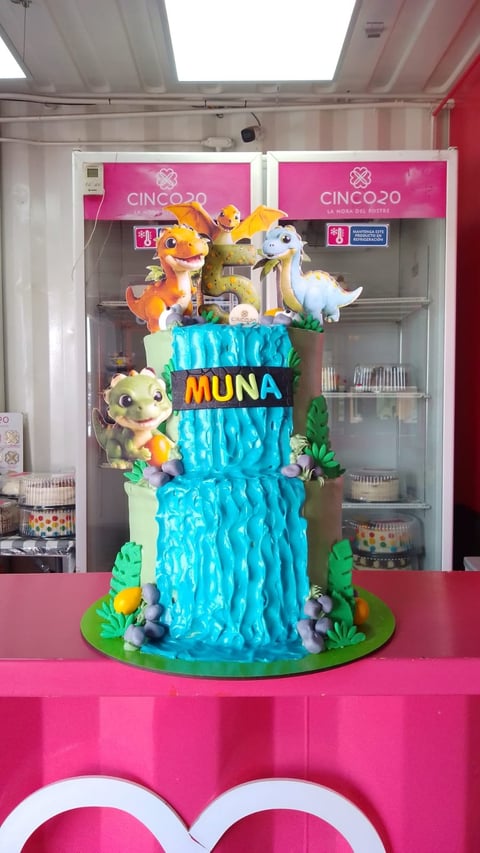 Three-tiered blue and white cake decorated with cartoon animal figures and MUNA text on pink table display